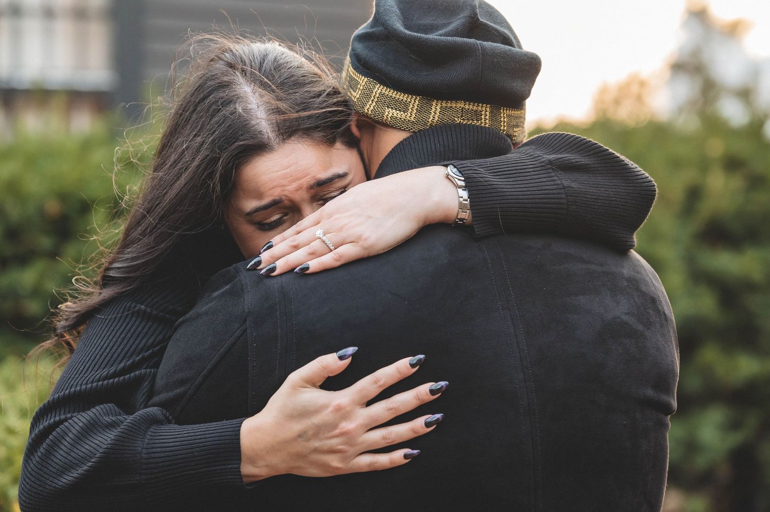 A tender moment captured as two people embrace in front of a grey house.