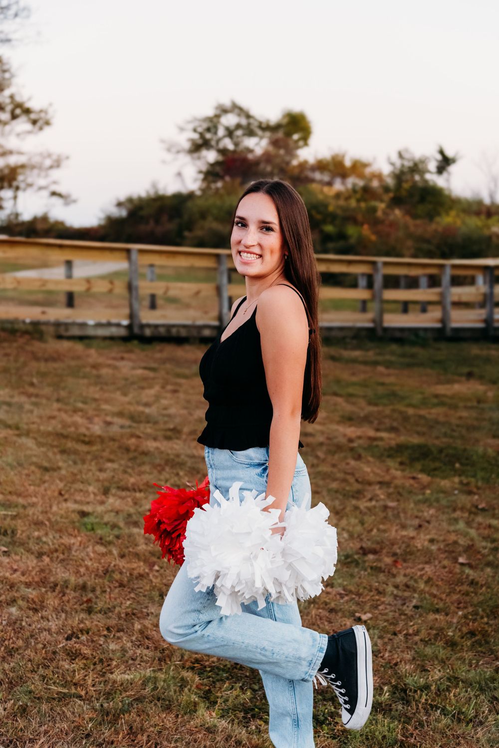 A cheerful person in light denim jeans and a black tank top poses outdoors with white and red pom poms near a wooden fence.
