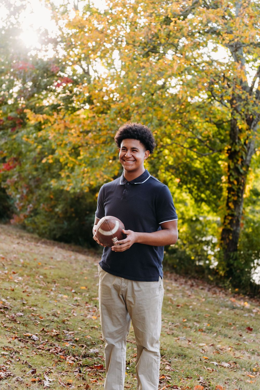 Someone in a black polo shirt holds a football while standing in a park with golden autumn foliage in the background.
