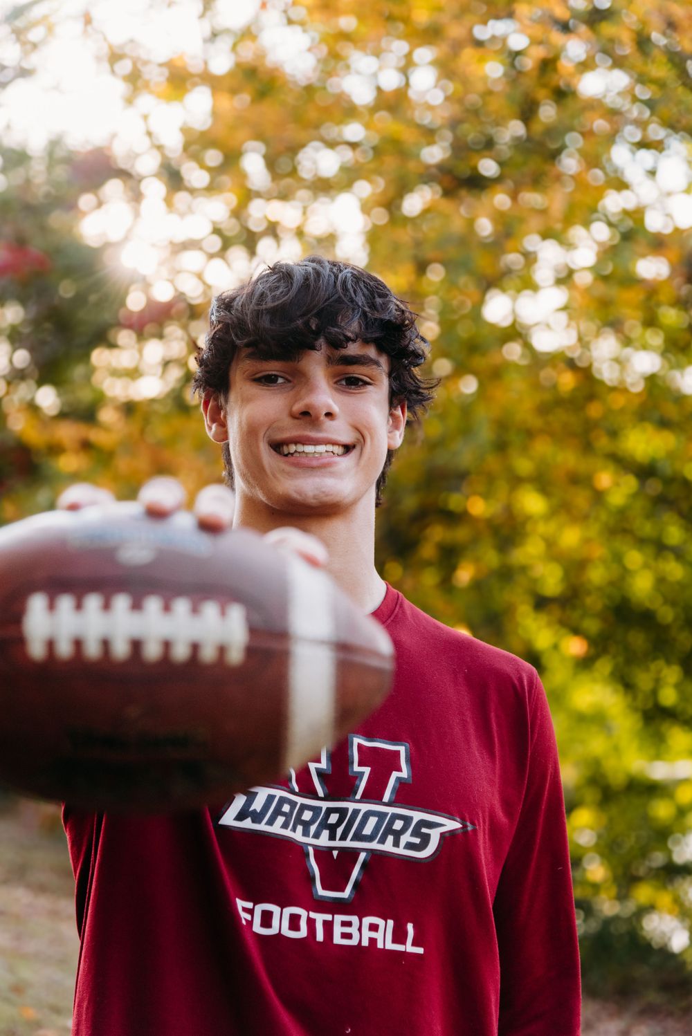 A football player in a red Warriors team sweatshirt holds up a football against an autumn backdrop with golden trees.