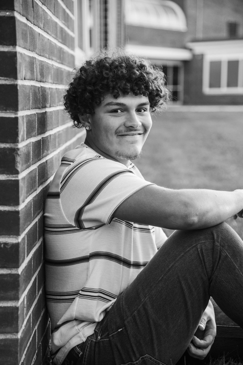 Black and white portrait of a young person with curly hair sitting against a brick wall and wearing a striped shirt.