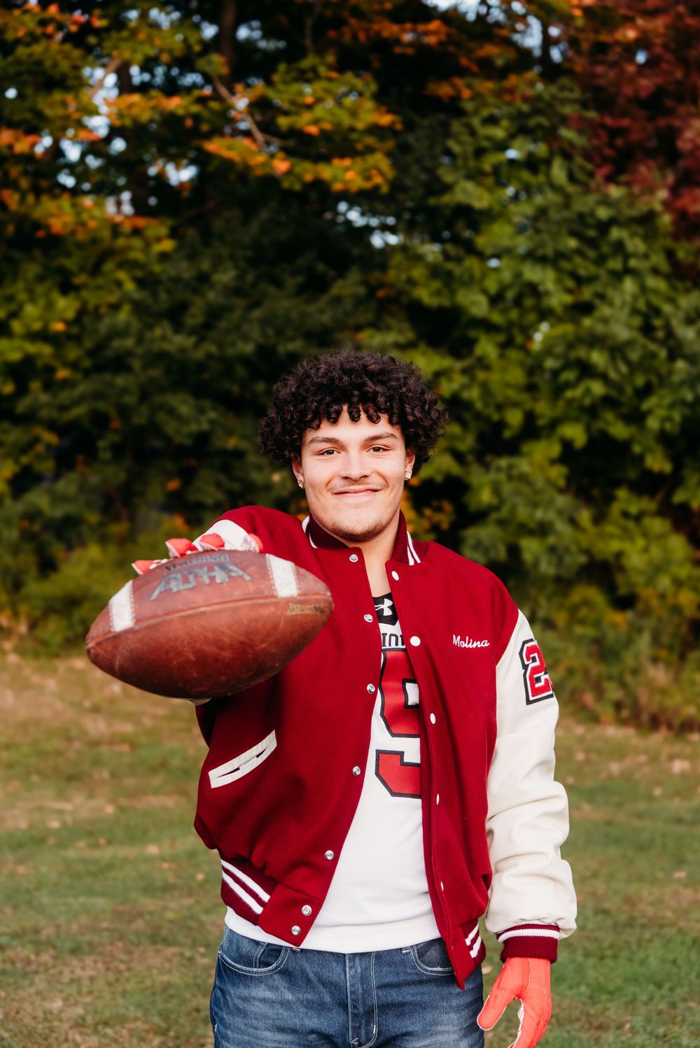 A student athlete in a red and white varsity jacket holds a football on an autumn day with fall foliage in background.