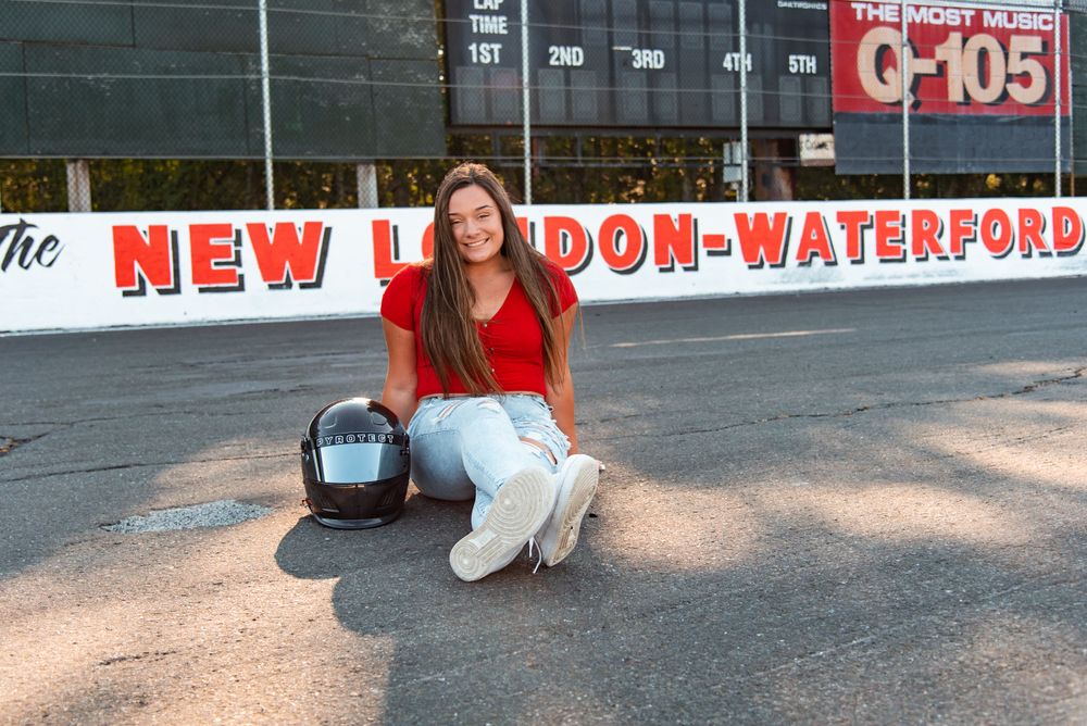 A person in a red sweater and white pants kneels on the asphalt at New London-Waterford Speedway.