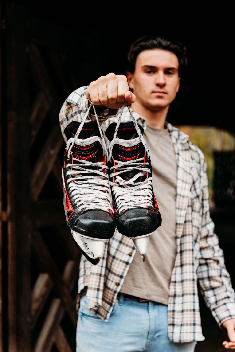 A person holding up worn hockey skates while wearing a plaid shirt and jeans in outdoor setting.