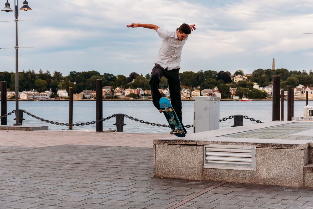 A skateboarder performs a stylish trick off a concrete ledge along a waterfront boardwalk during daytime.