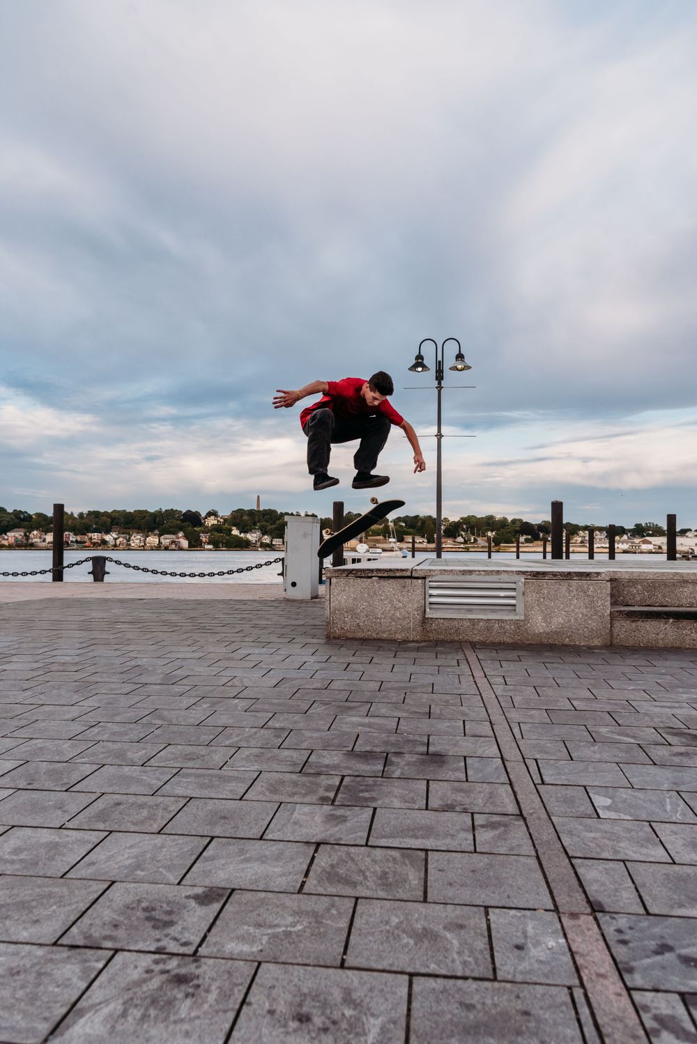 Skateboarder in red performs an aerial trick over a concrete ledge at a waterfront plaza under cloudy skies.