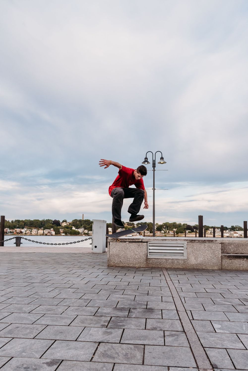 A skateboarder performing an ollie trick over a concrete barrier at a waterfront plaza under cloudy skies.
