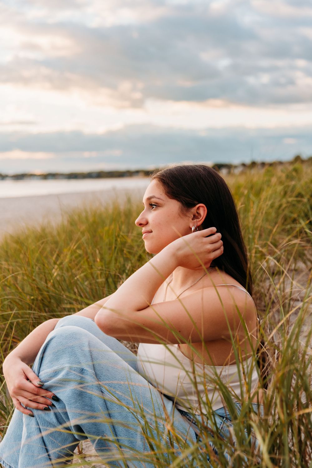 Person sits thoughtfully in tall beach grass at sunset beside a tranquil waterfront.