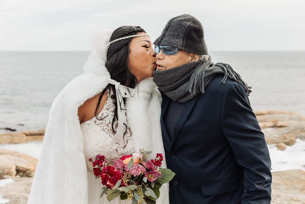 A romantic winter wedding couple embraces on a rocky beach shore holding a burgundy and pink bouquet in cold weather.