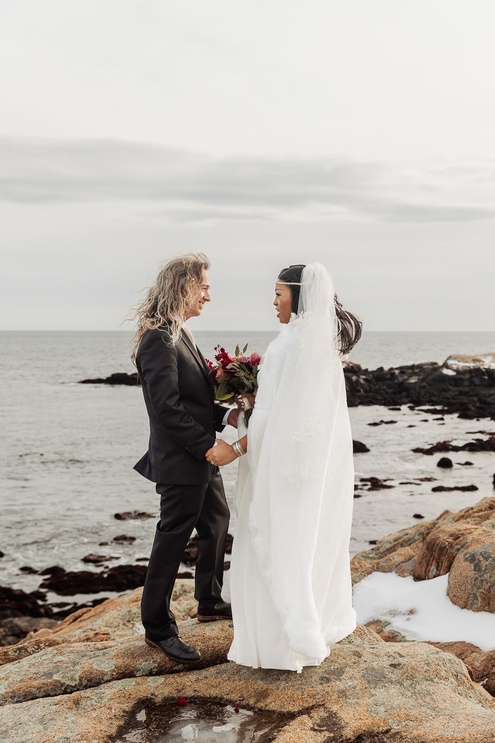 A couple shares an intimate moment during their wedding ceremony on rocky coastal cliffs overlooking a moody ocean.