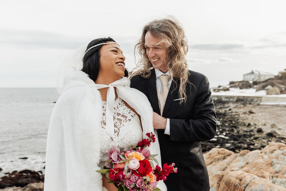 A couple shares a joyful moment together on a rocky coastal beach, with one wearing a white cape and holding a red bouquet.