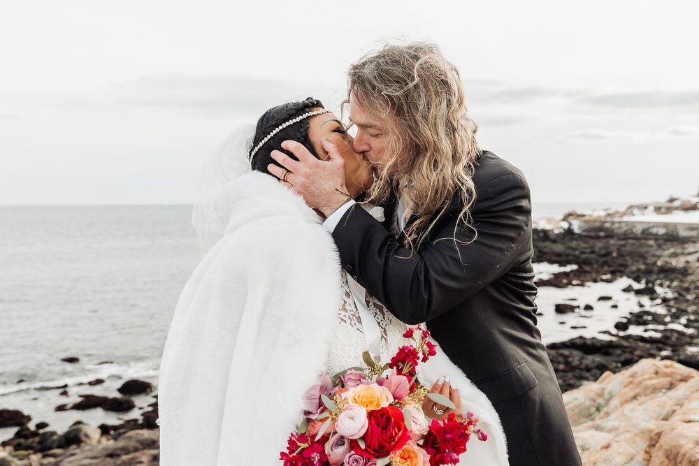 A loving couple shares a romantic moment at a rocky beach with vibrant red floral bouquet in hand.