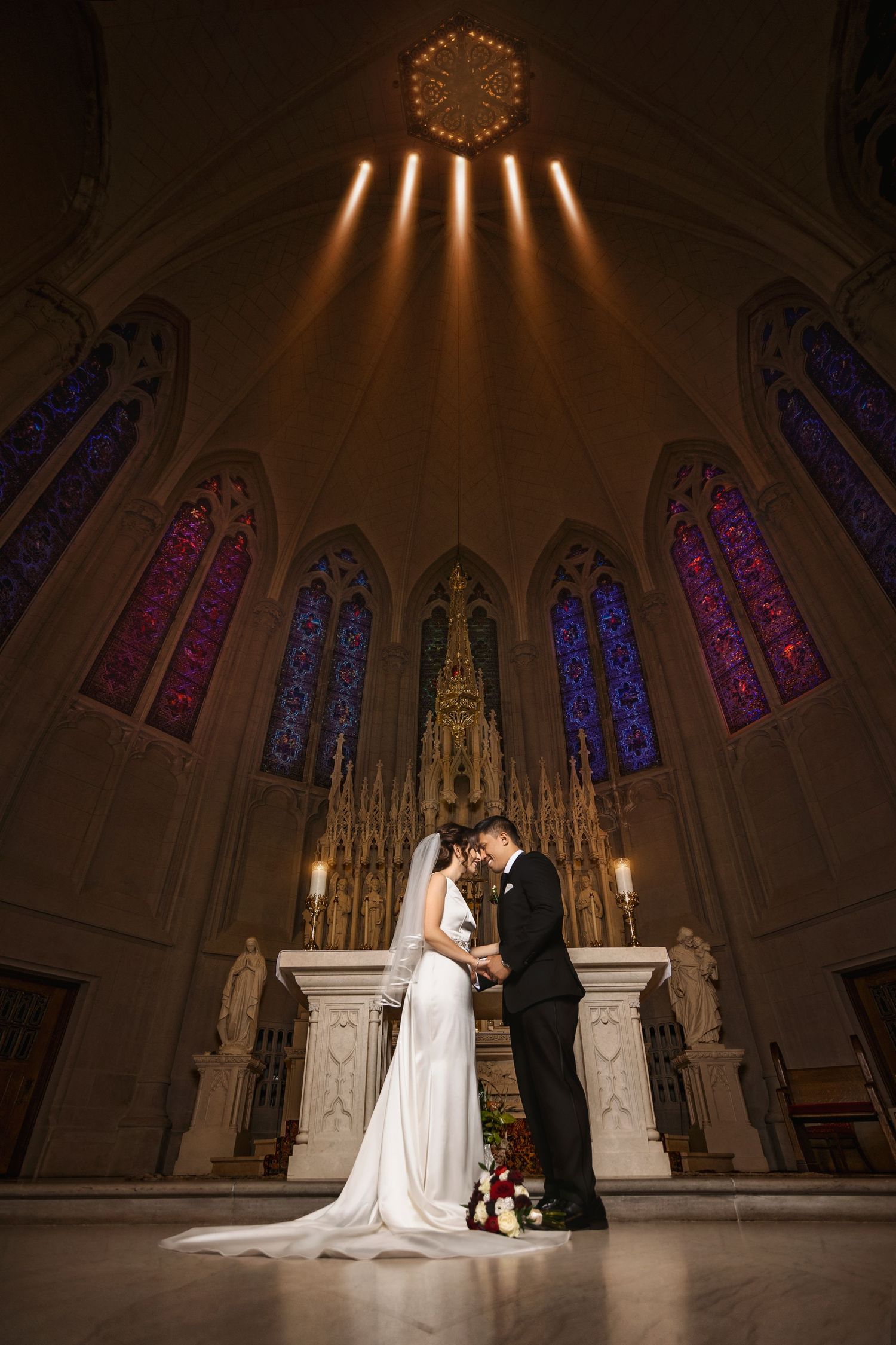 A couple embraces at a wedding ceremony in a gothic cathedral with dramatic lighting and stained glass windows.