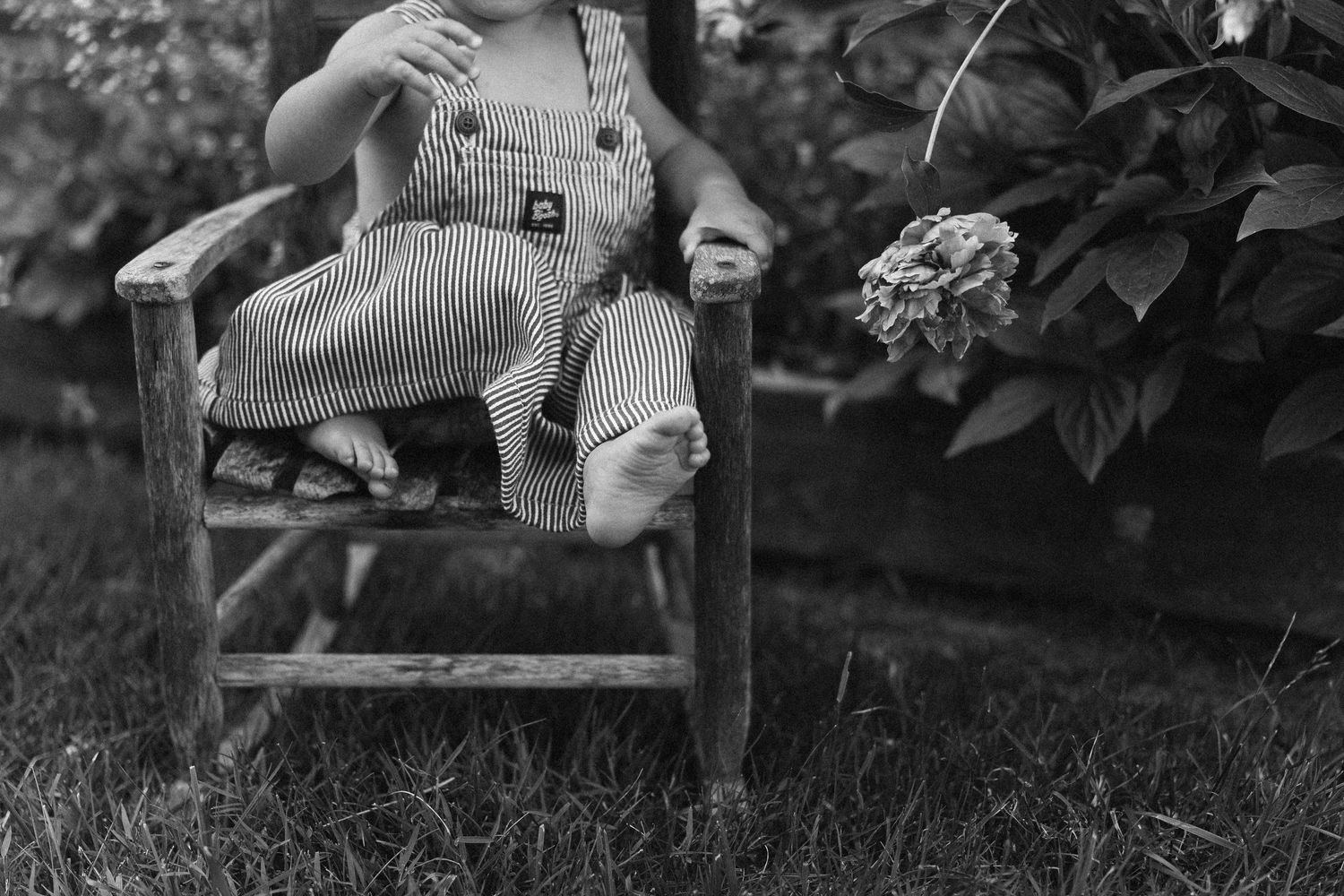 Vintage black and white photograph of small child in overalls sitting on rustic wooden chair in garden setting.