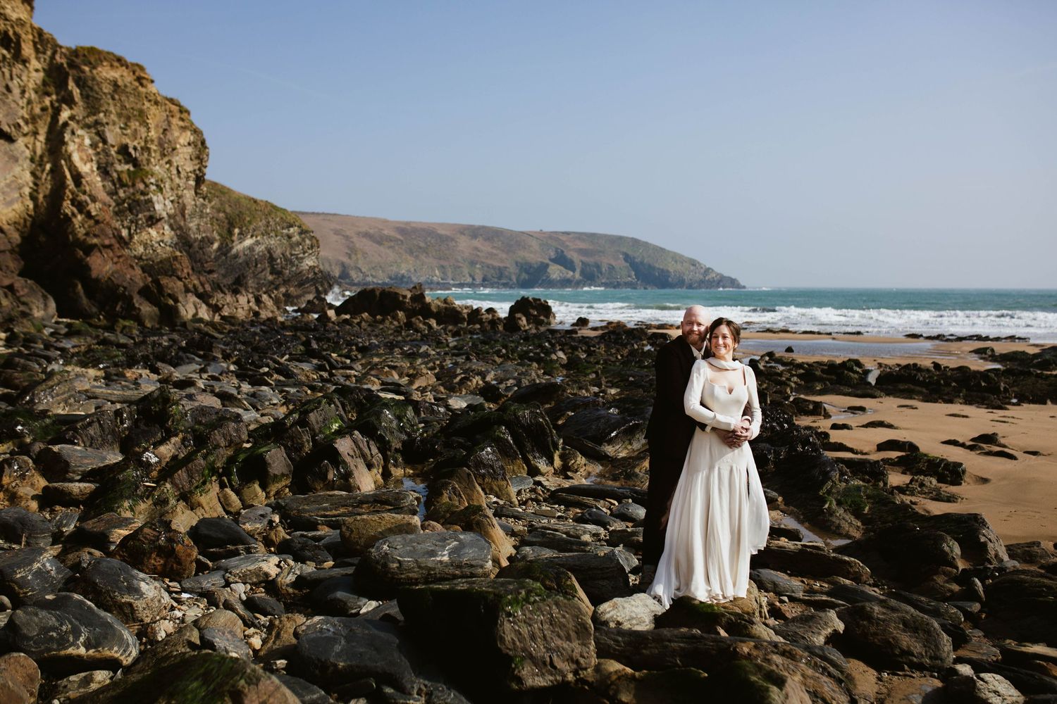 Stunning coastal wedding celebration on a rugged beach with dramatic cliffs and rocky shoreline in the background.