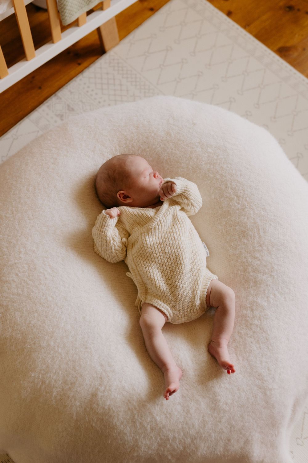 A small infant wearing a cream-colored knit romper lies sleeping on a plush white cushion with hardwood flooring visible.