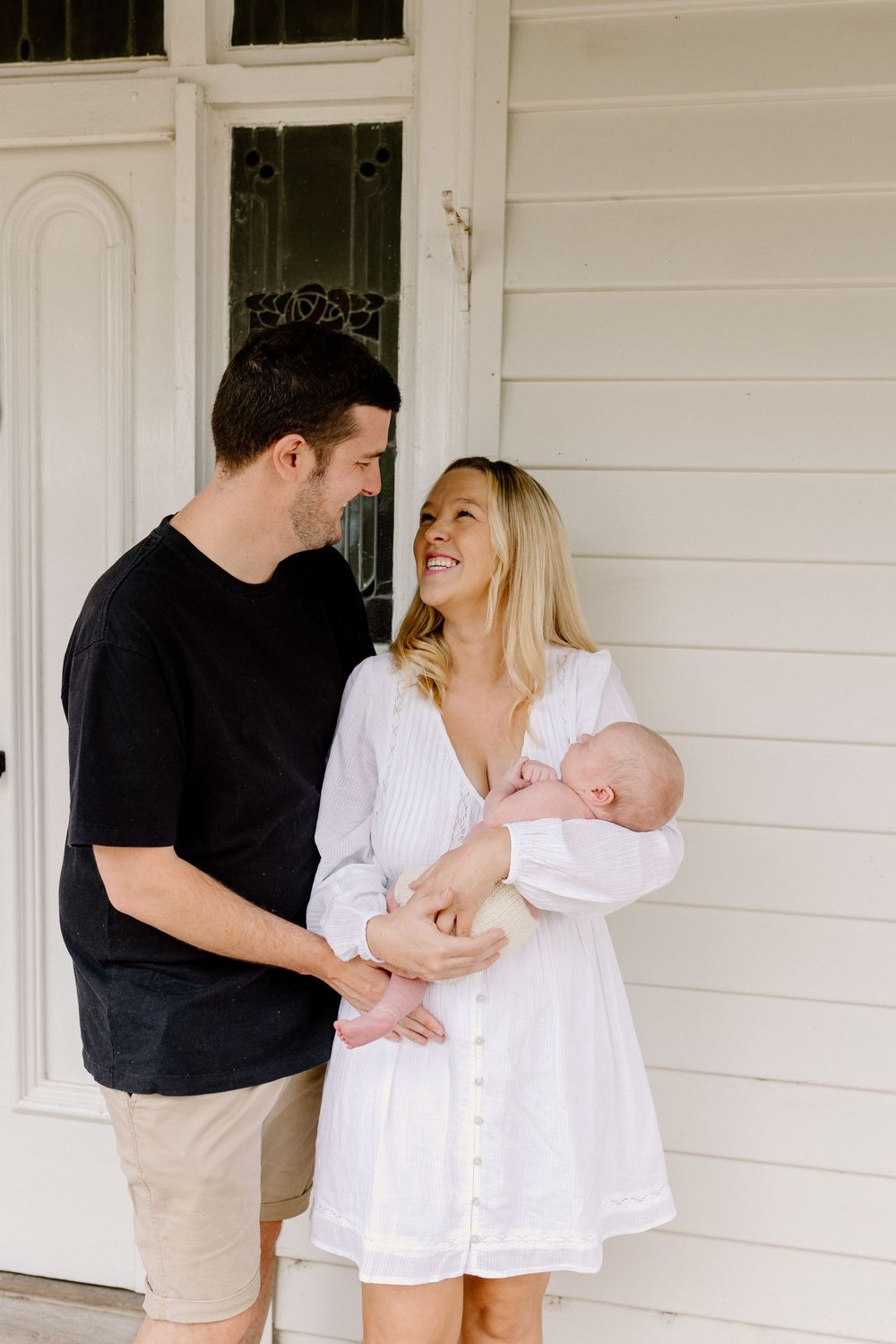 A couple in black and white attire stand together on a porch with a baby in a sweet candid moment.