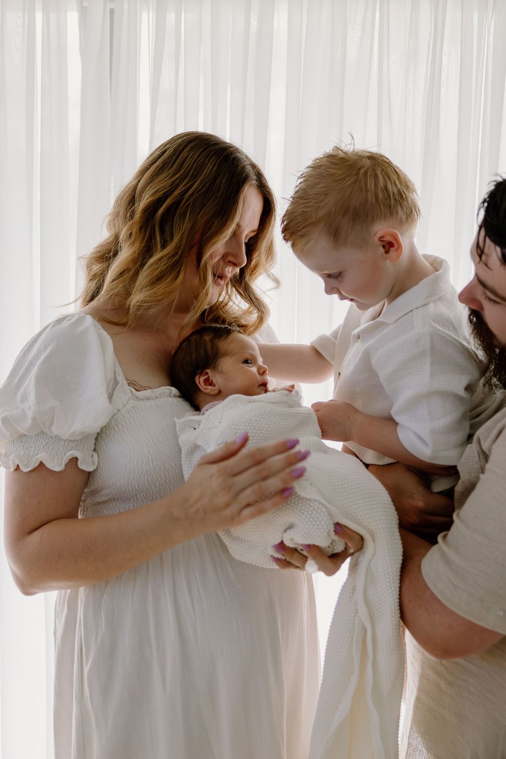 Family in white clothing sharing an intimate moment together as they gently interact with a newborn baby.