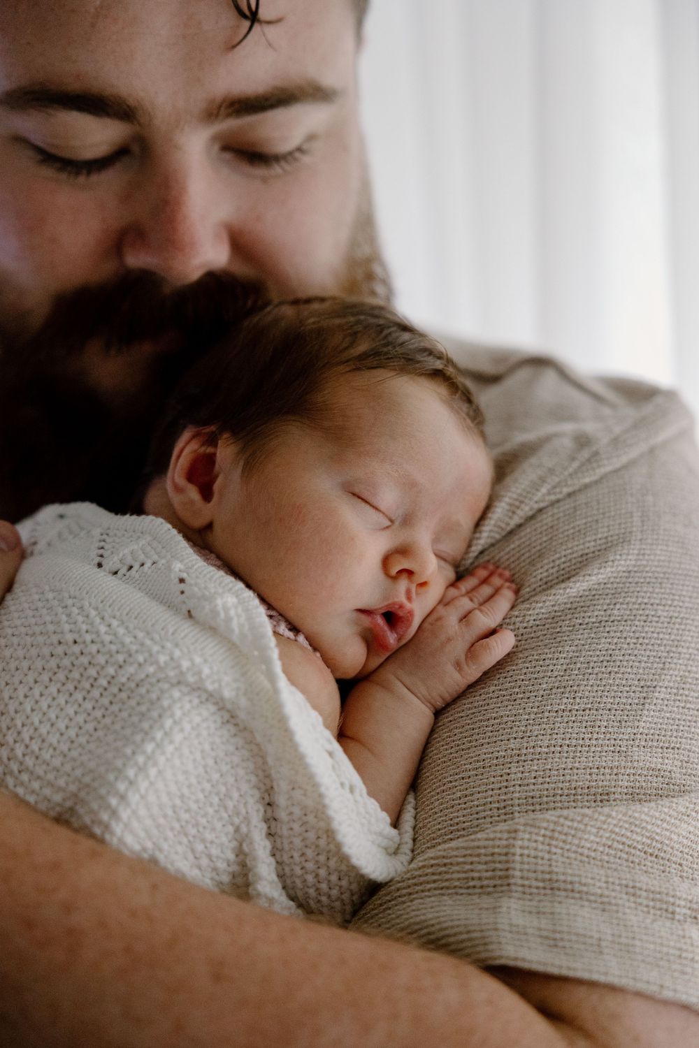 A bearded person tenderly holds a sleeping infant wrapped in a cozy white knit blanket against their chest.