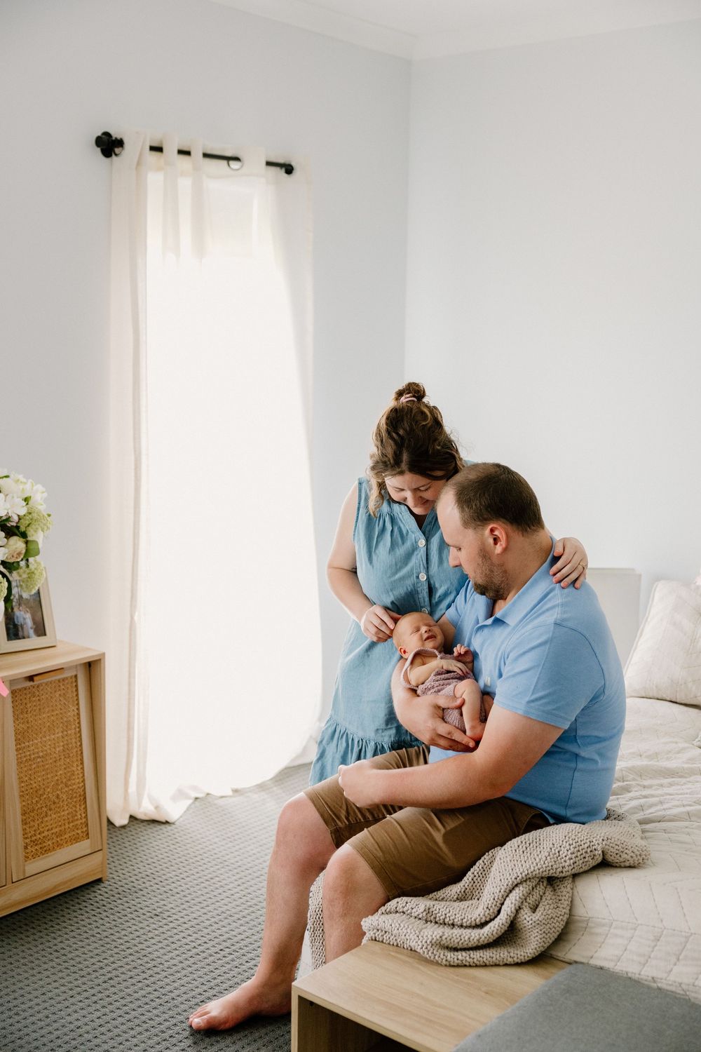 A couple sits together in bright room holding their newborn baby while dressed in casual blue and brown outfits.