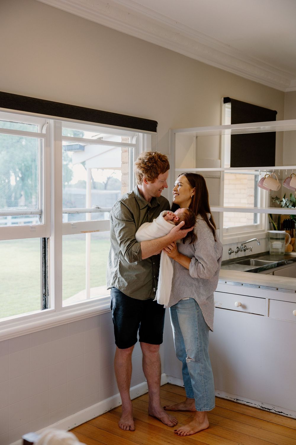 Two people standing barefoot in a bright kitchen with large windows sharing an intimate moment together.
