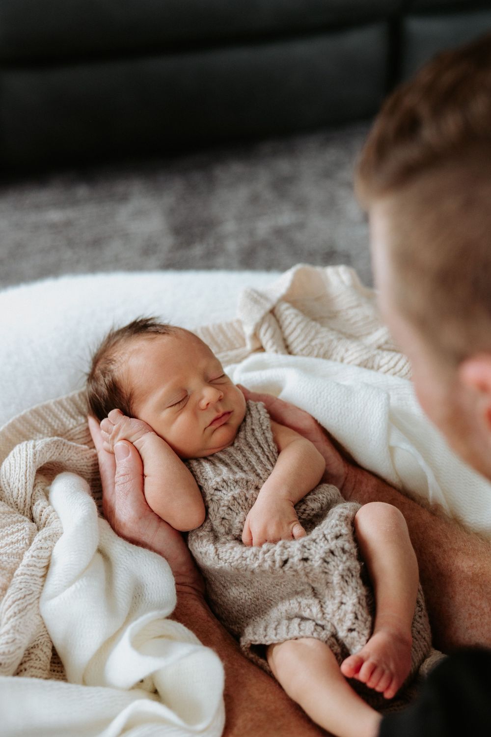 A sleeping newborn baby wearing a knitted romper lies peacefully on soft white blankets.