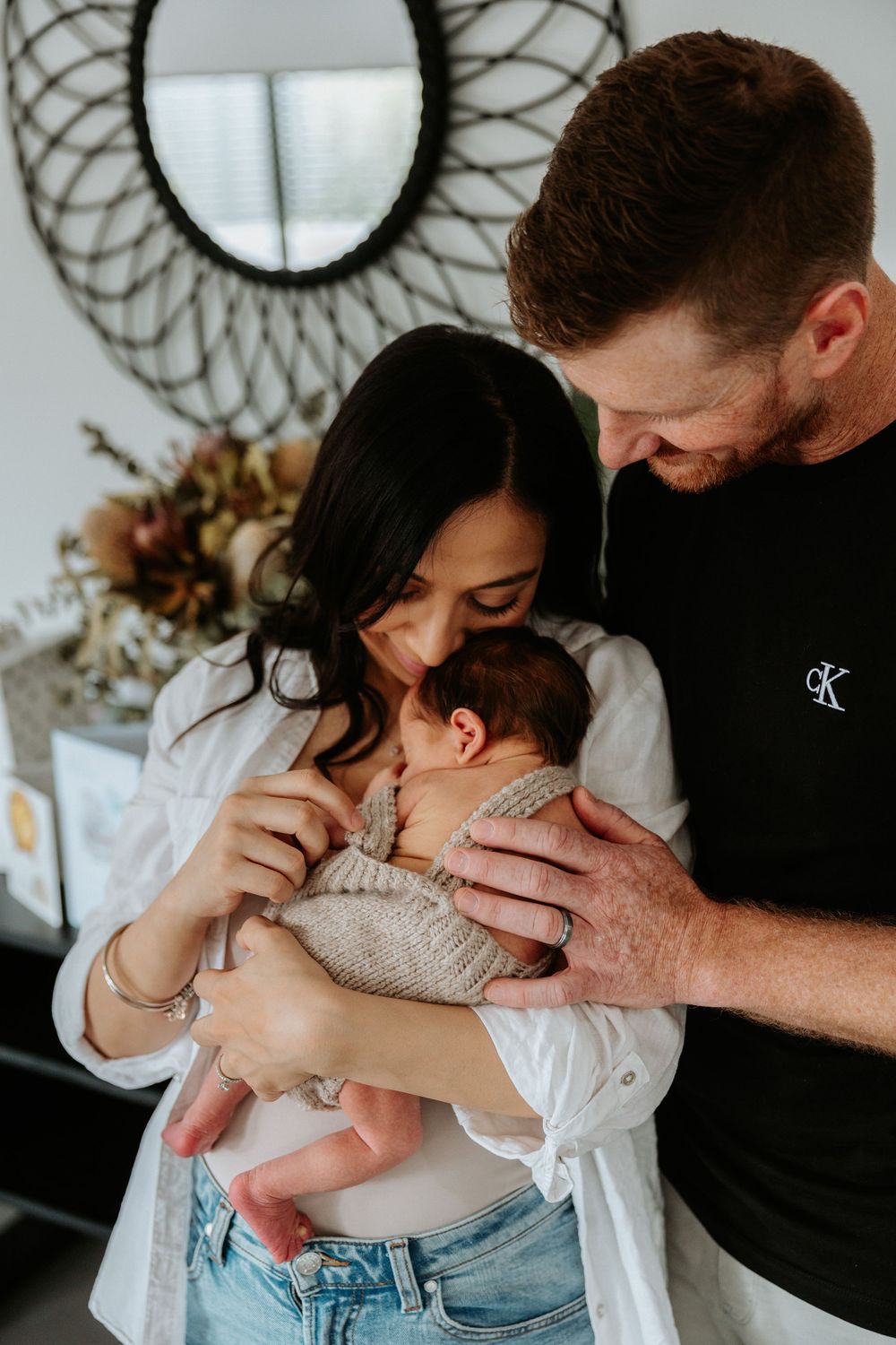 A couple sharing an intimate moment with their newborn baby beneath a modern sunburst mirror.