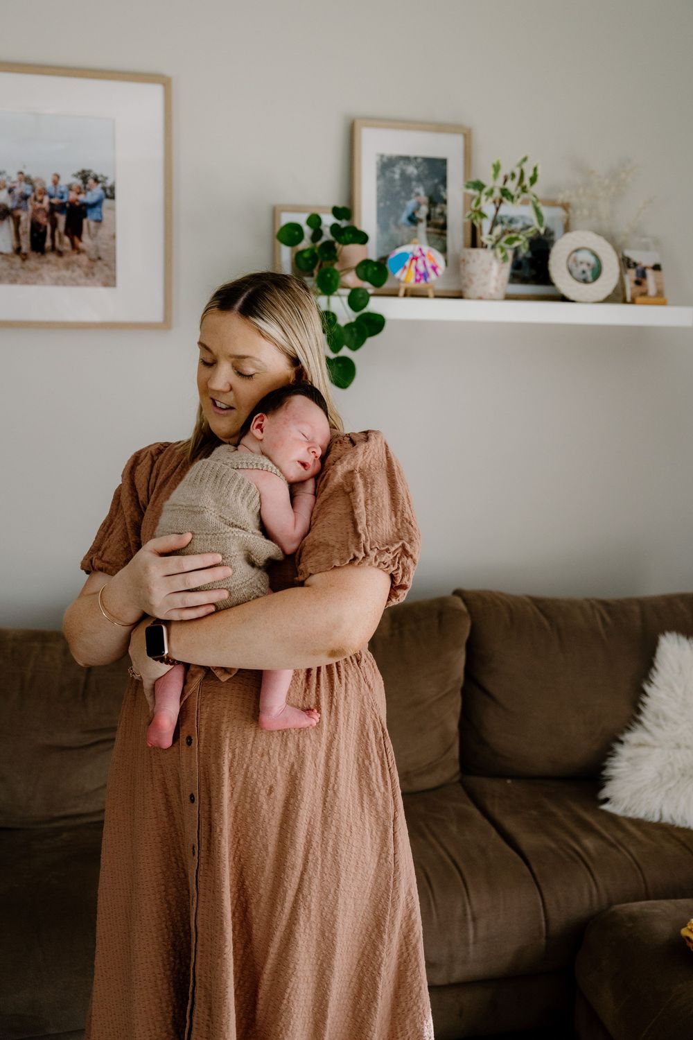 A caregiver in a brown dress stands in a cozy living room adorned with plants and photographs, holding a sleeping baby.