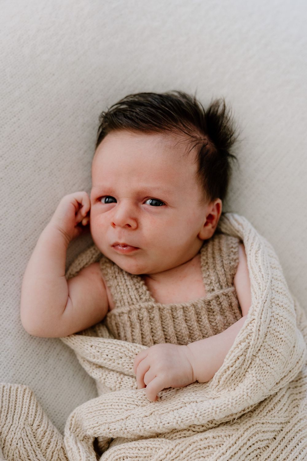 A small baby lying on their side wearing a cozy knit sweater against a beige background.