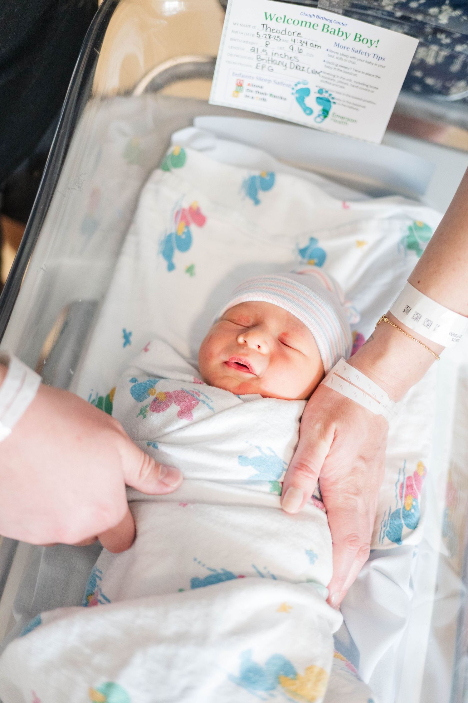 A newborn baby wrapped in patterned blanket sleeps peacefully in hospital bassinet with welcome card visible above.