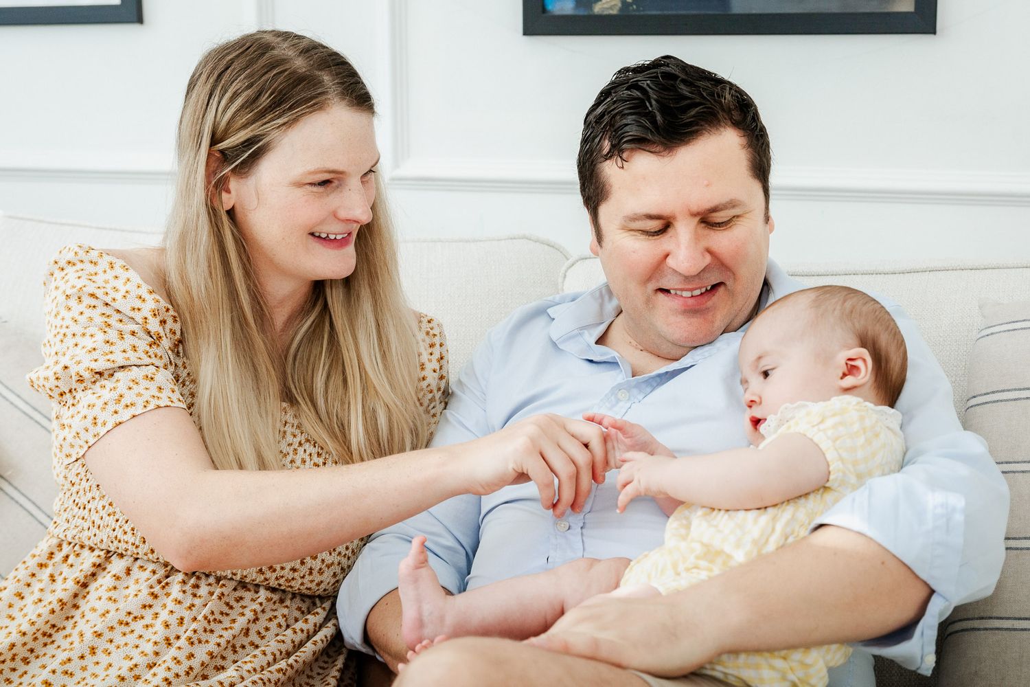 A mum and dad cuddle their baby on the sofa at home. They smile and look relaxed. Bristol motherhood photography by Little Bodhi Photography