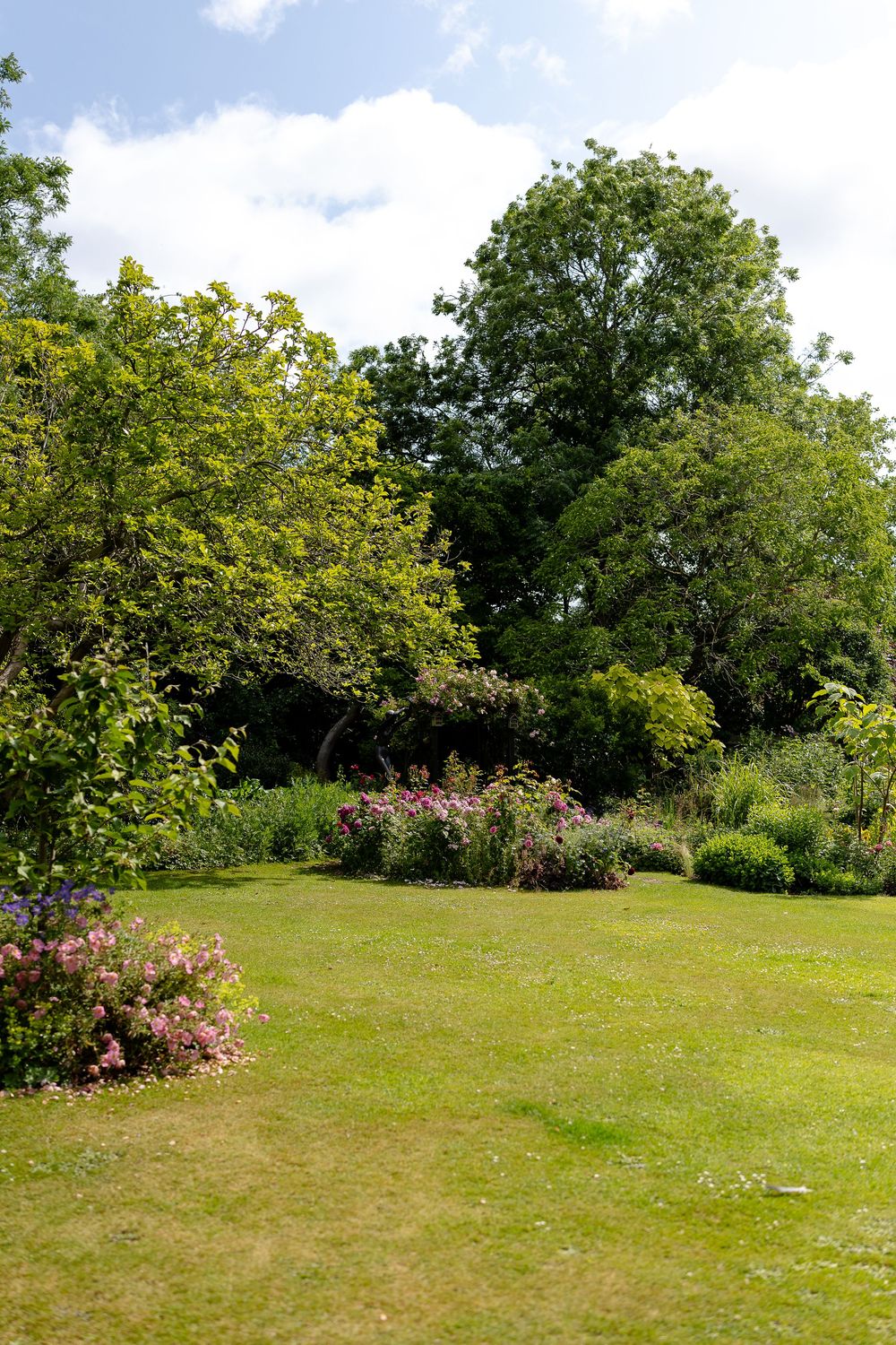 Scenic view of a lush green garden lawn with blooming flower bushes and mature trees under a partly cloudy summer sky.