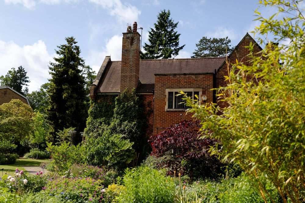 A rustic brick house with tall evergreen trees and lush gardens surrounds the property on a sunny day.