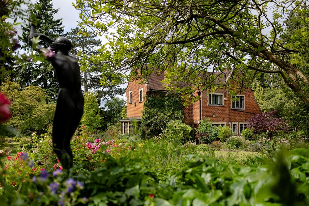 Garden art stands among colorful roses and greenery with a red brick house in the background.