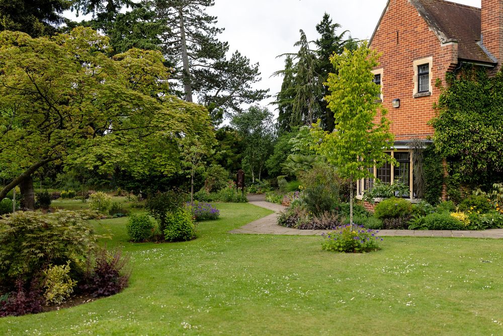 A well-maintained garden landscape with green lawn, flowering beds and a red brick house surrounded by mature trees.