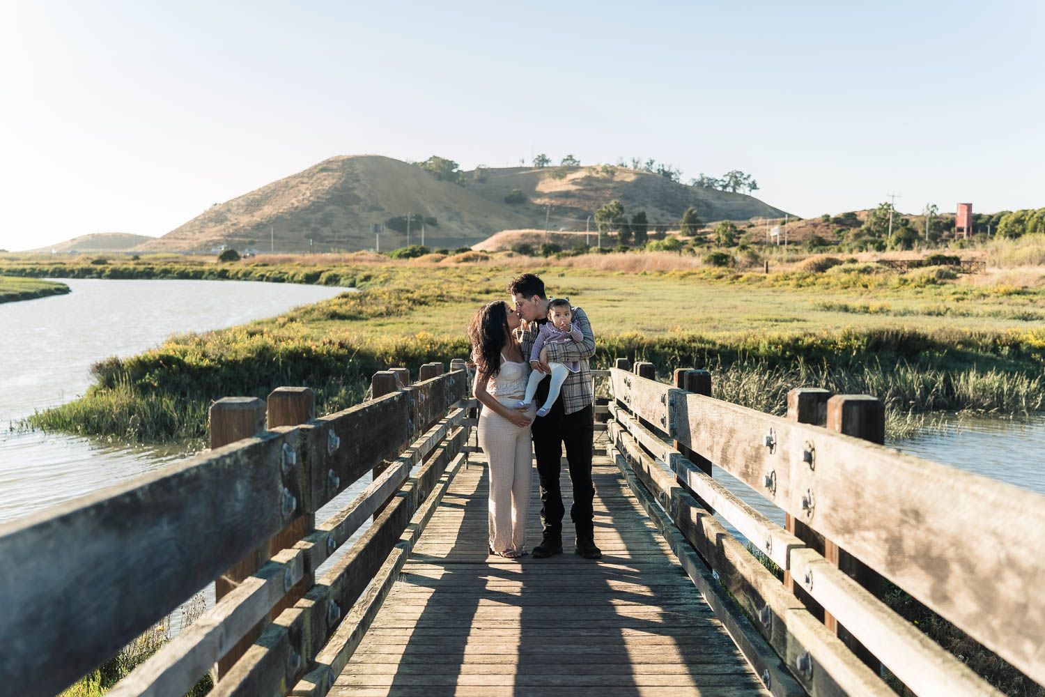 Family with a pregnant woman and young girl standing on a wooden boardwalk over marshland with rolling hills in the background.