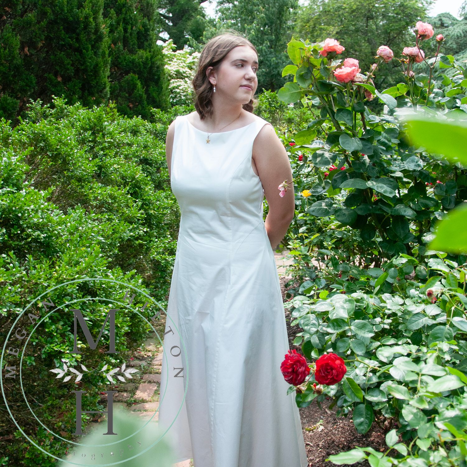 A sleeveless white dress worn among blooming red roses and lush garden greenery on a summer day.