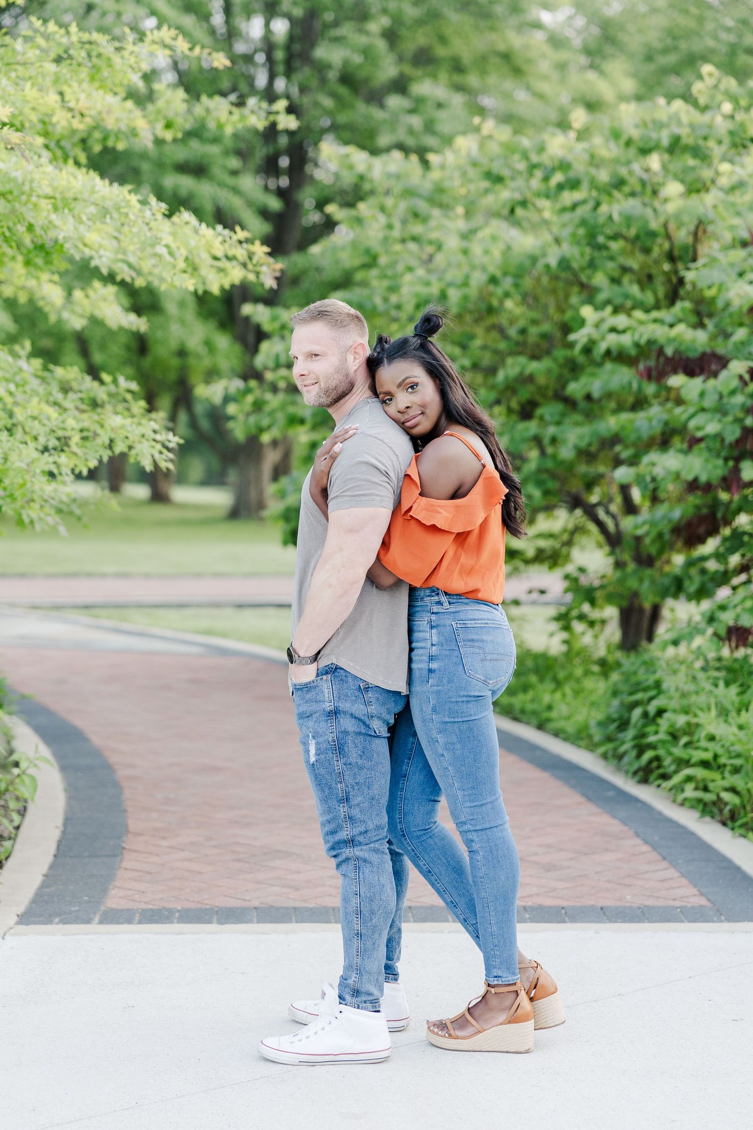 Couple stands back-to-back on a brick pathway surrounded by lush green foliage in a park setting.