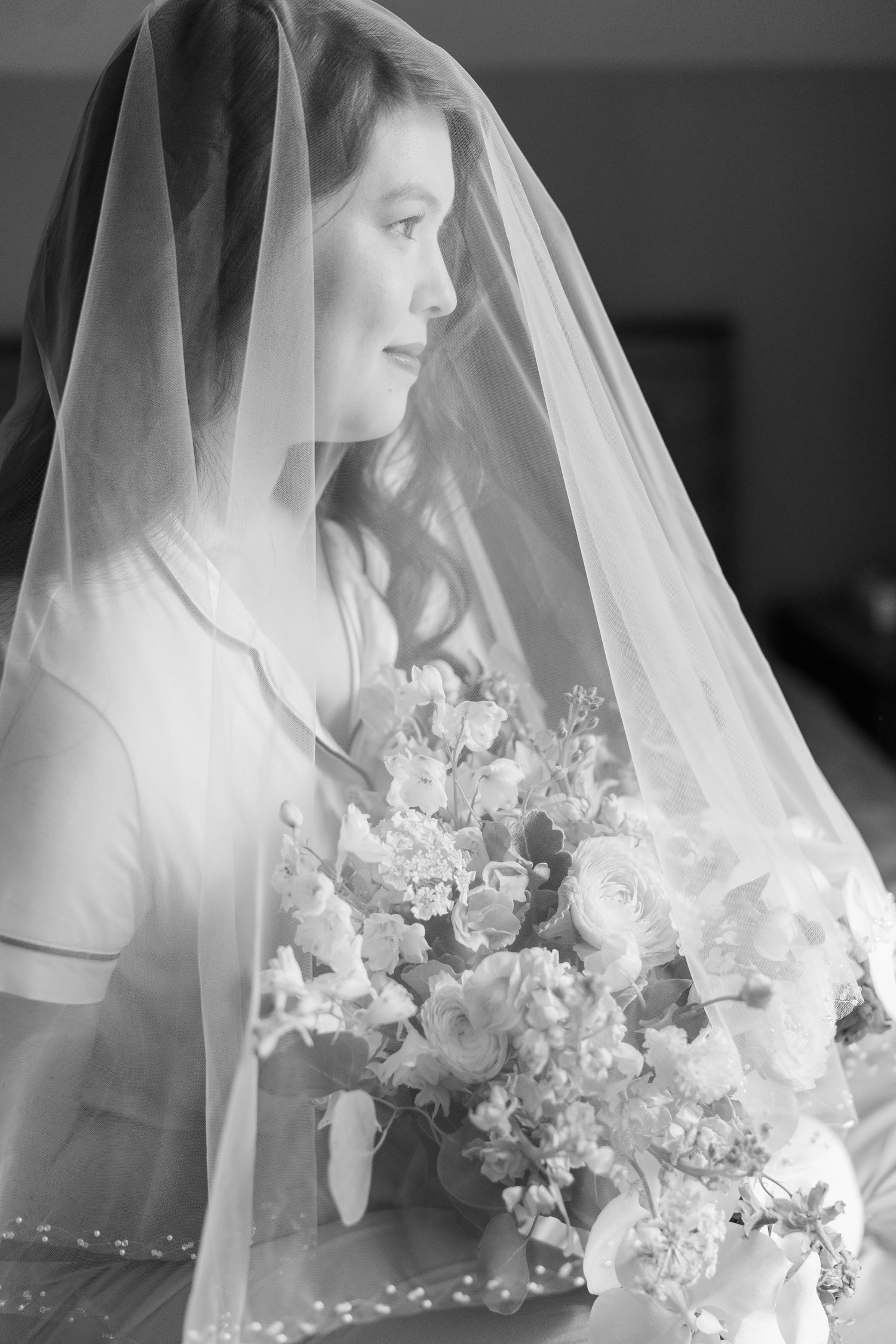 A bride in a delicate veil holds a lush bouquet of white flowers while posing in an elegant black and white portrait.