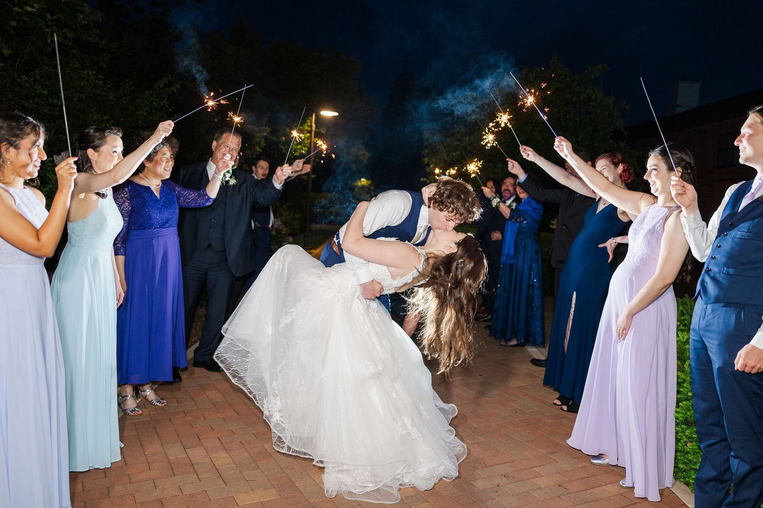 Wedding guests hold sparklers in a nighttime celebration as couple shares romantic dance on brick patio.