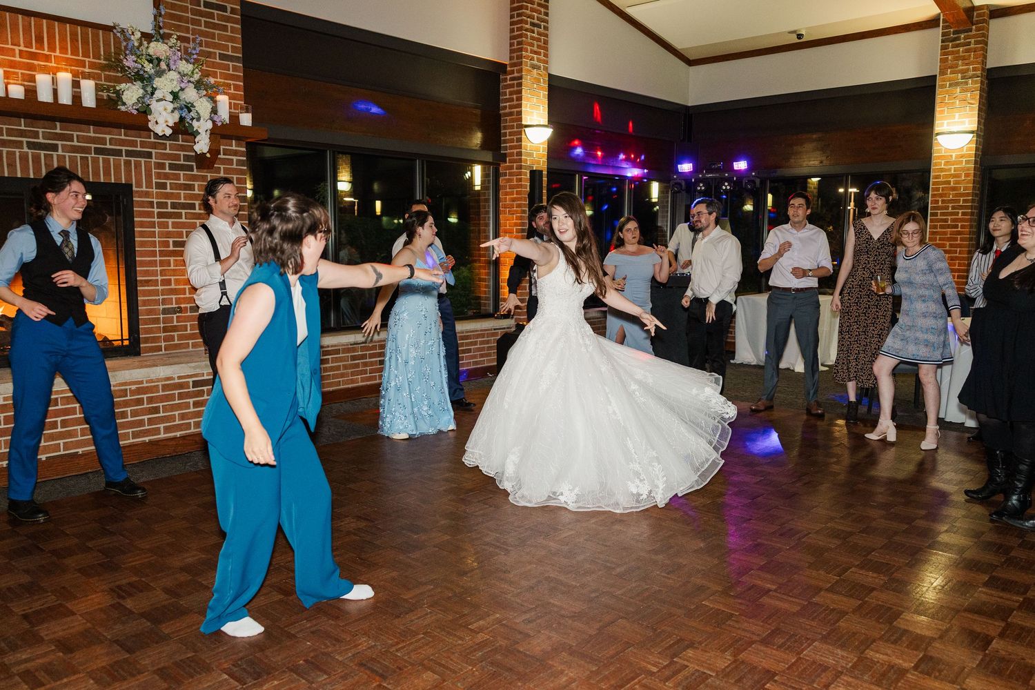 Party guests dancing on a wooden dance floor at a wedding reception with exposed brick walls and ambient lighting.