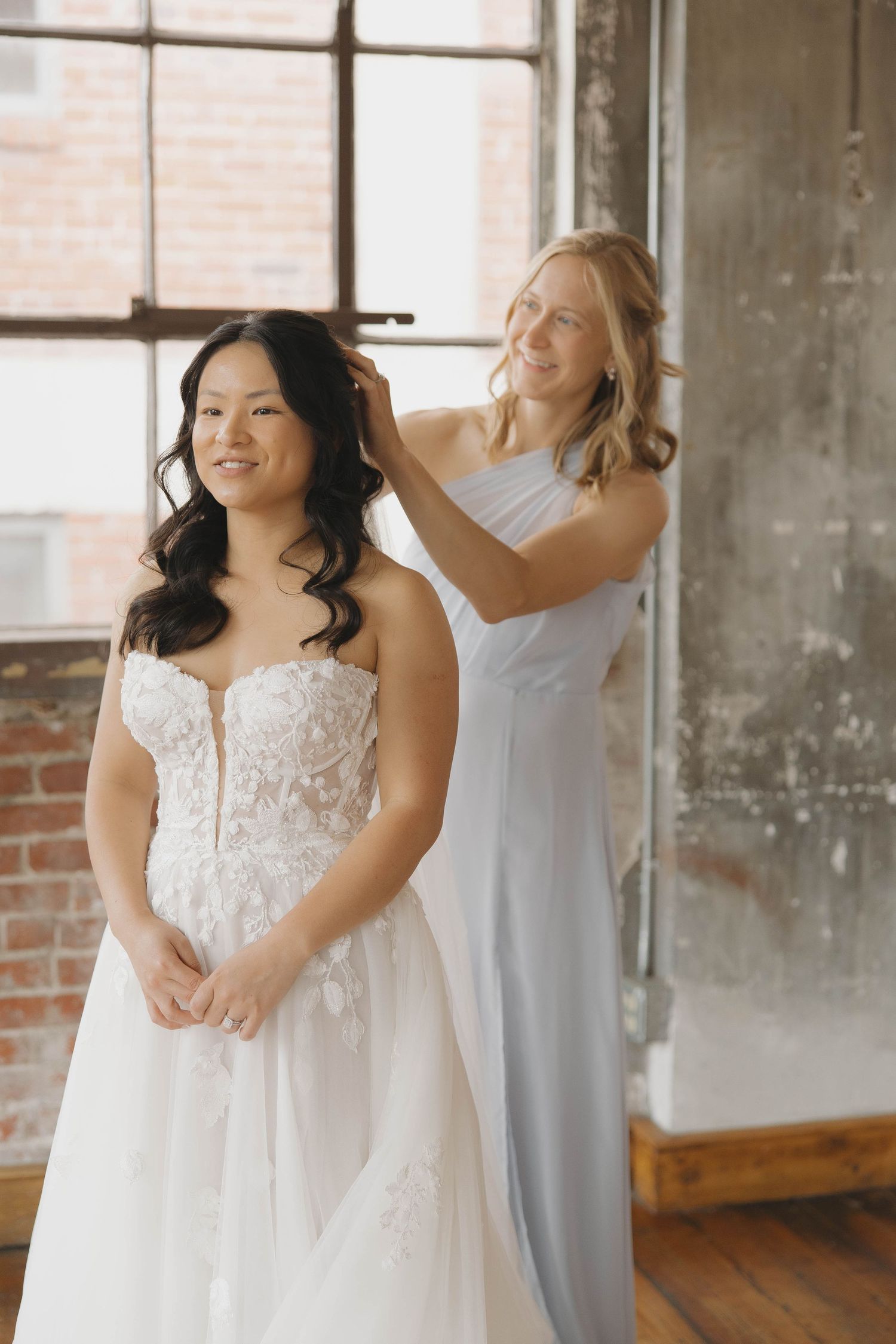 An elegant bridal preparation moment shows a person in a strapless white lace gown being assisted in a rustic room.