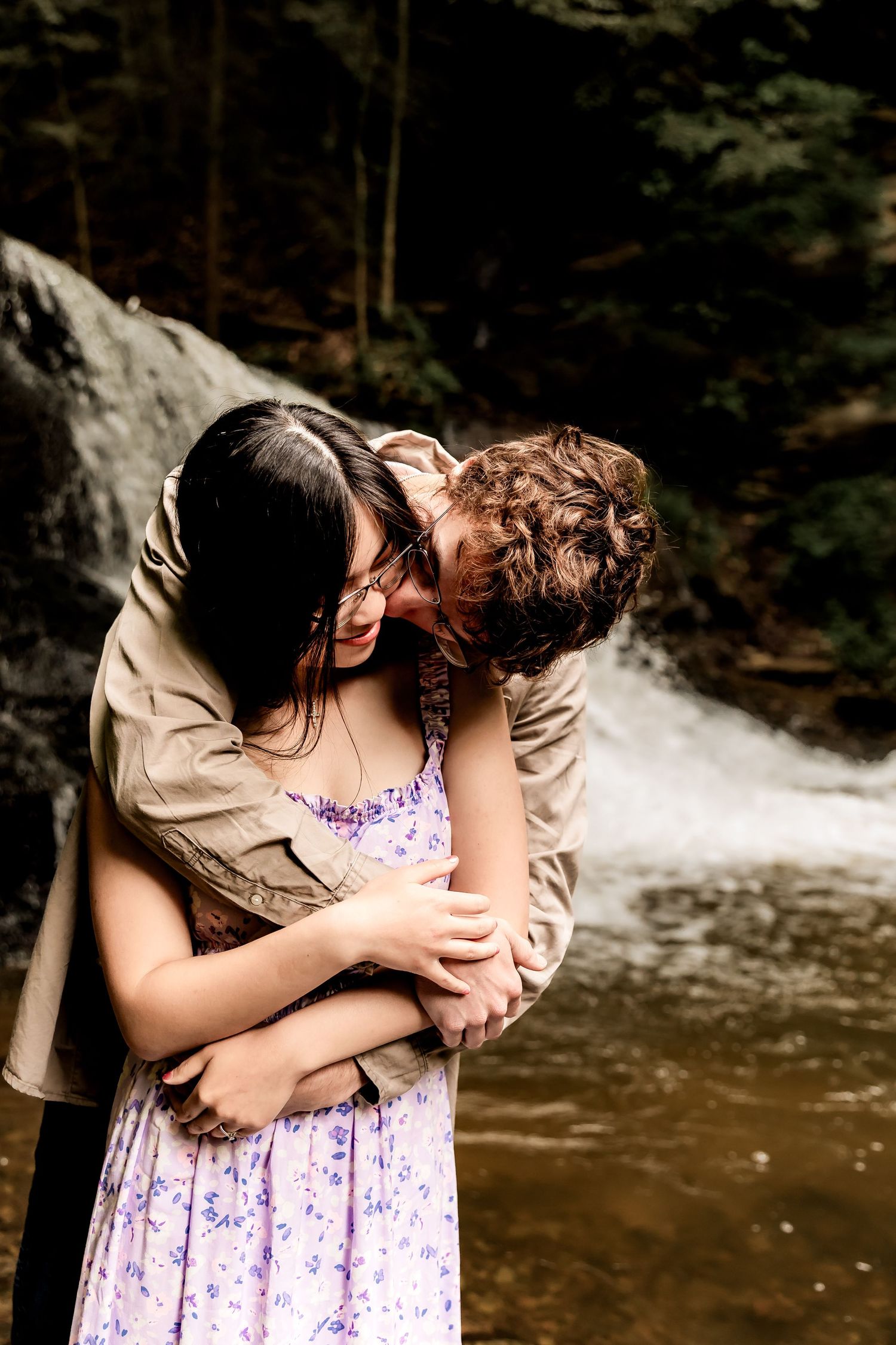 A romantic couple embraces by a forest waterfall, with one wearing a beige jacket and the other in a purple floral dress.