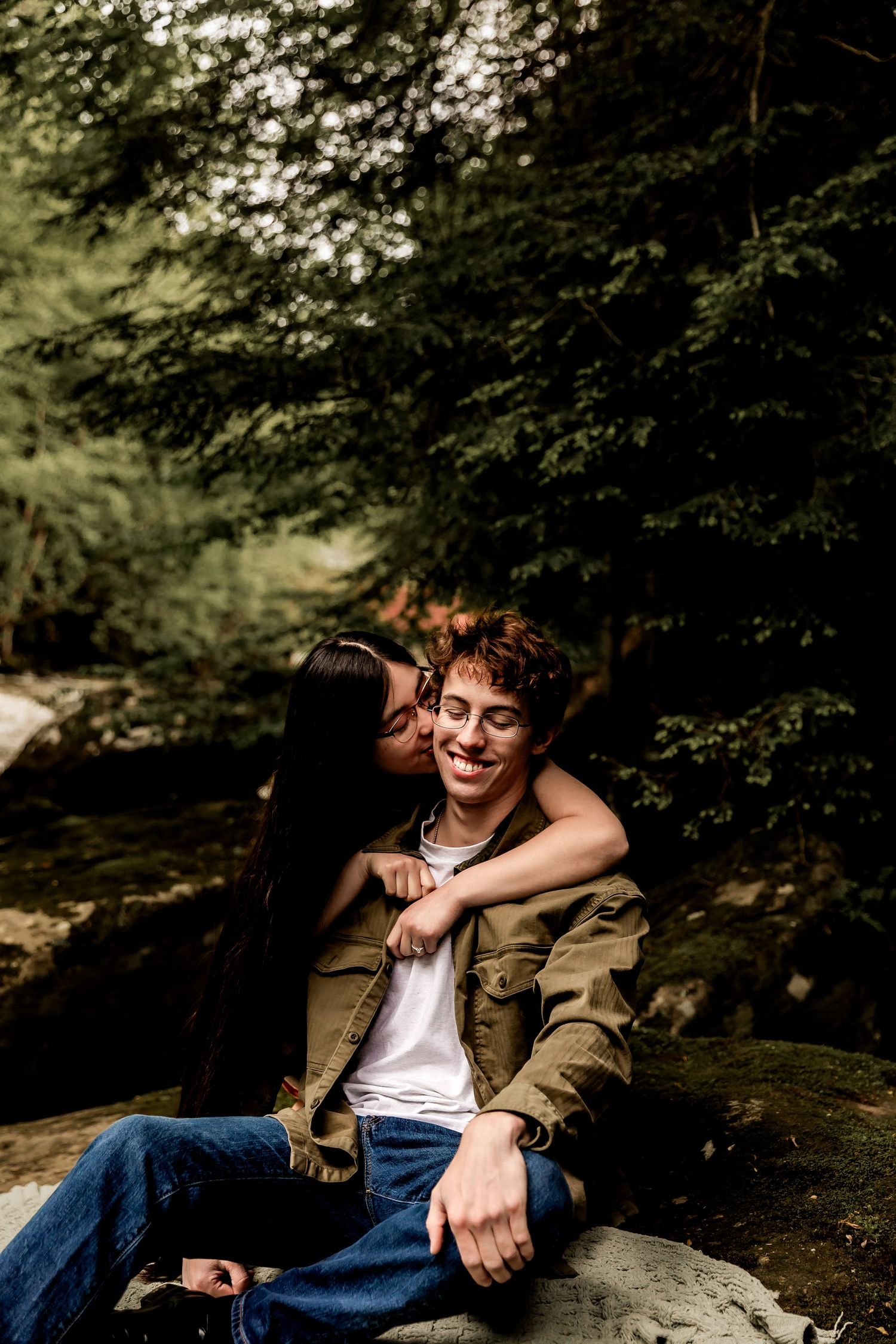 A person in an army green jacket and jeans sits relaxed on mossy rocks near a flowing stream in a wooded setting.