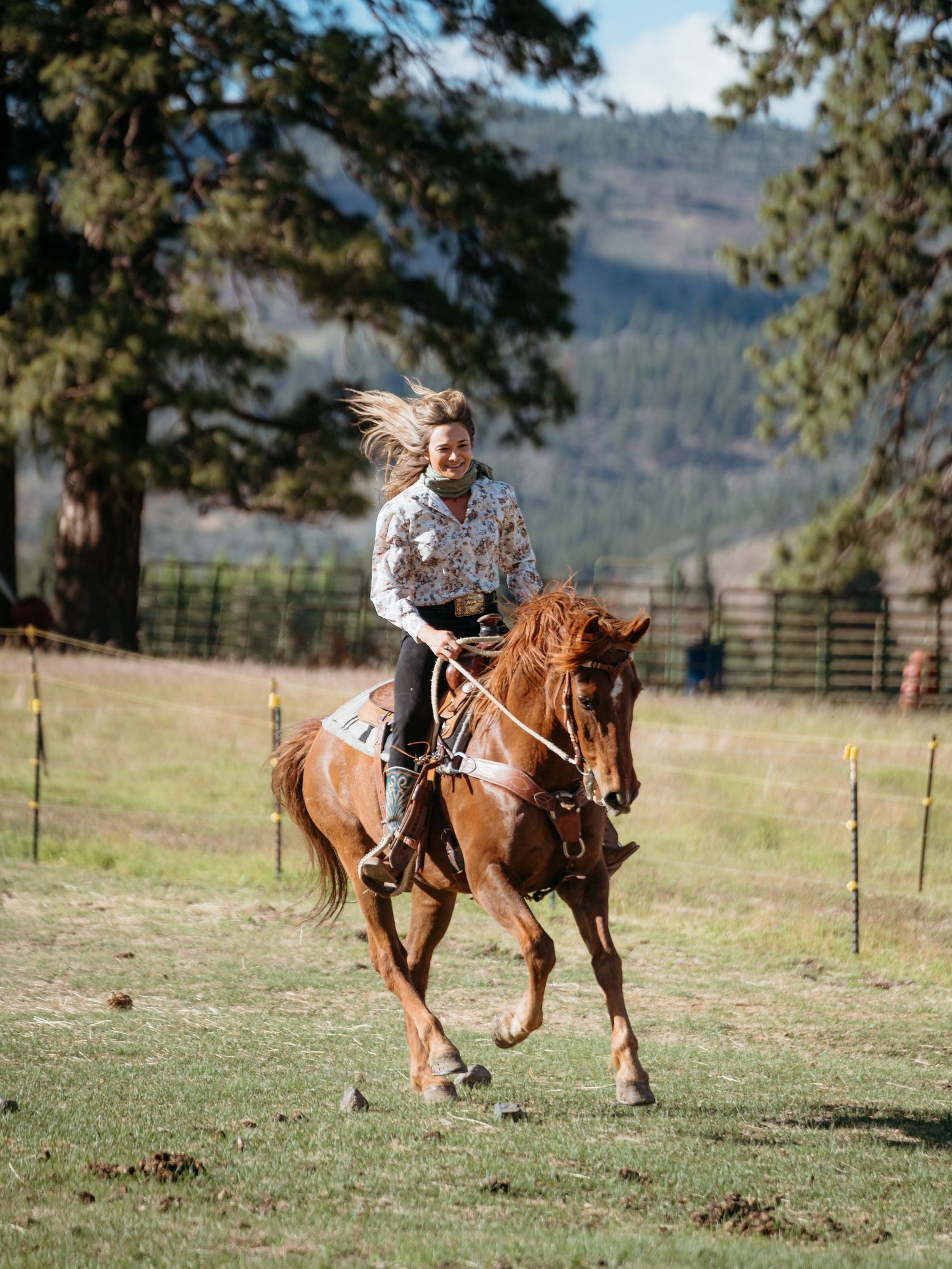 A rider on a chestnut horse gallops across a scenic mountain ranch with trees and hills in the background.