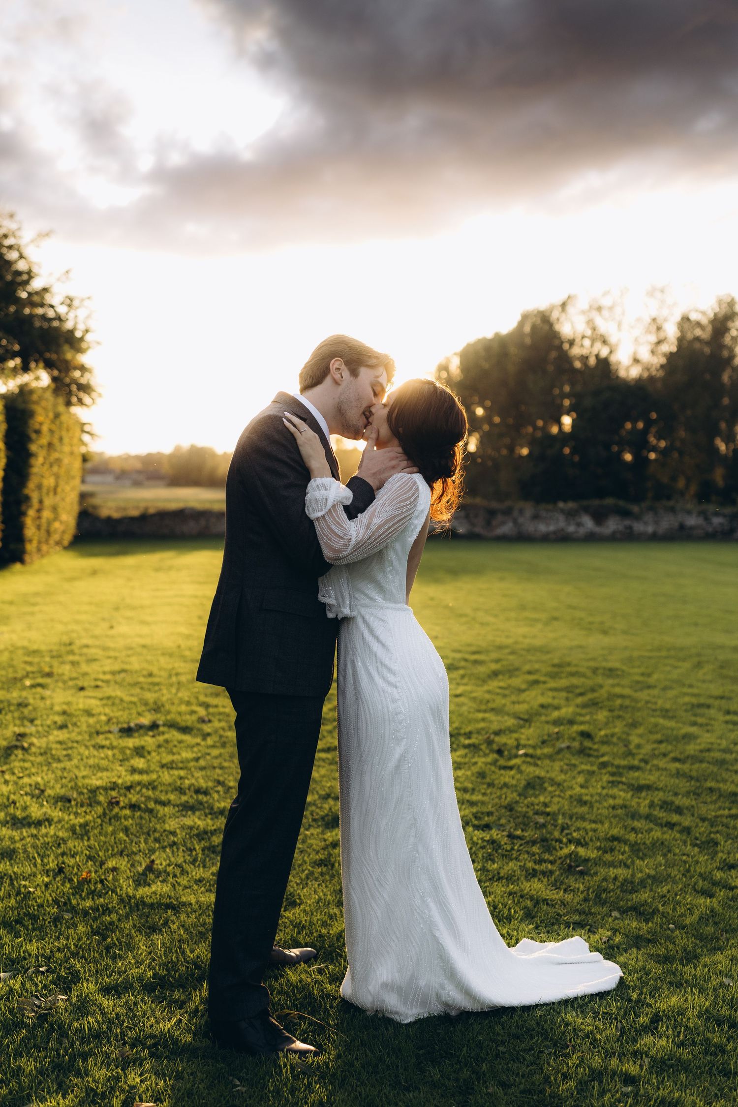A romantic sunset wedding photo of a couple embracing on a lush green lawn with golden light filtering through trees.