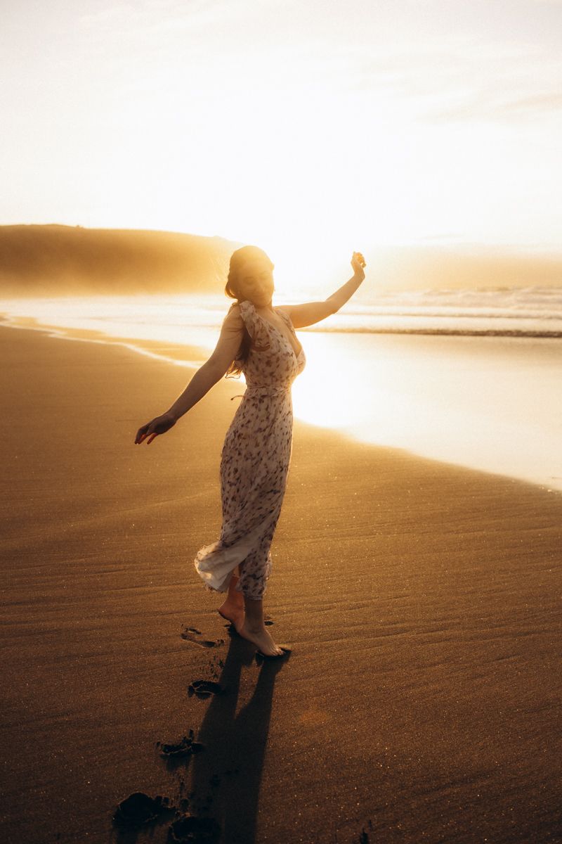 Silhouette of person in flowing dress dancing on golden beach at sunset casting long shadow on sand.