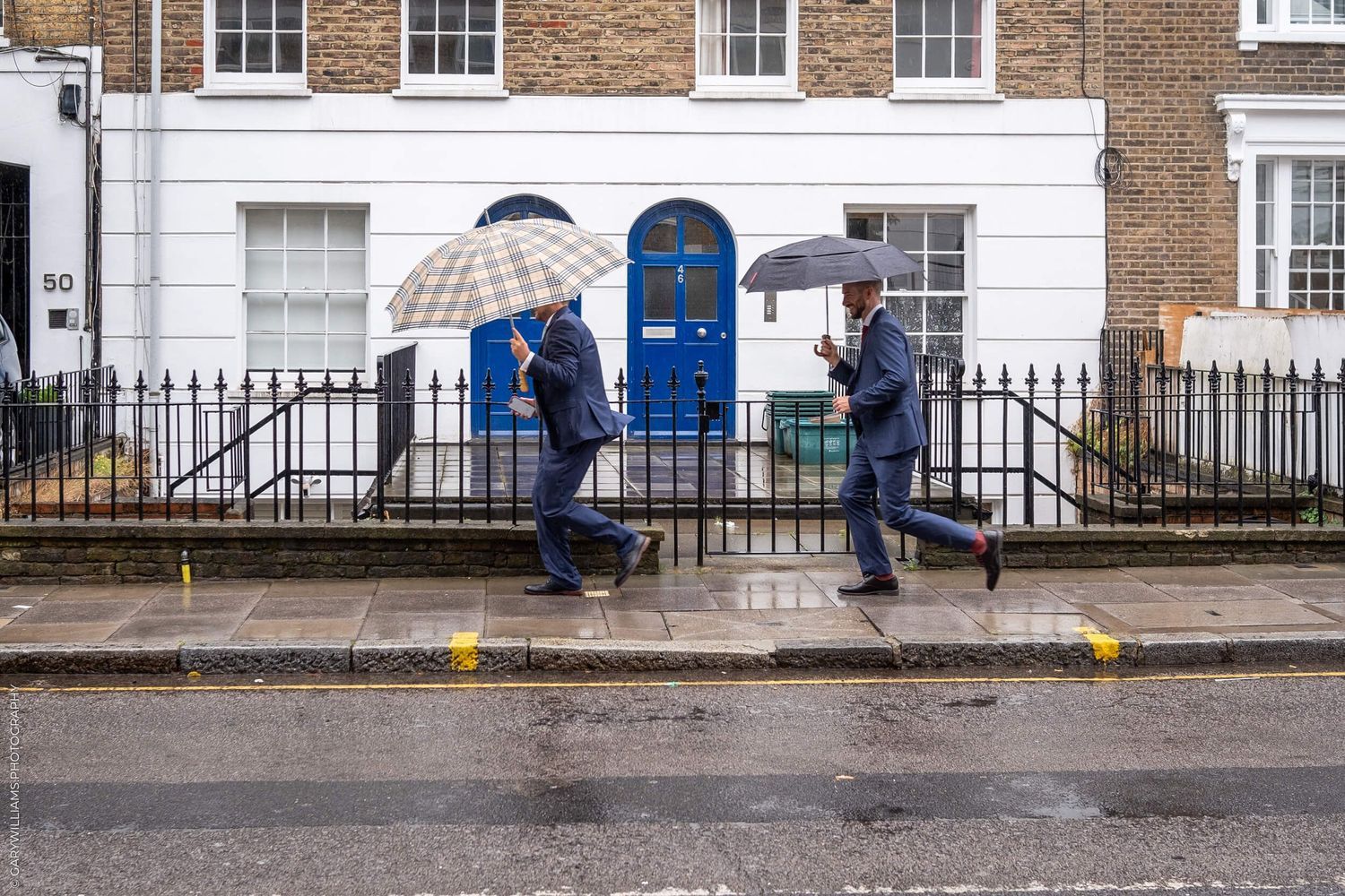 People walking with umbrellas on a rainy day past brick townhouses with a bright blue door in London.