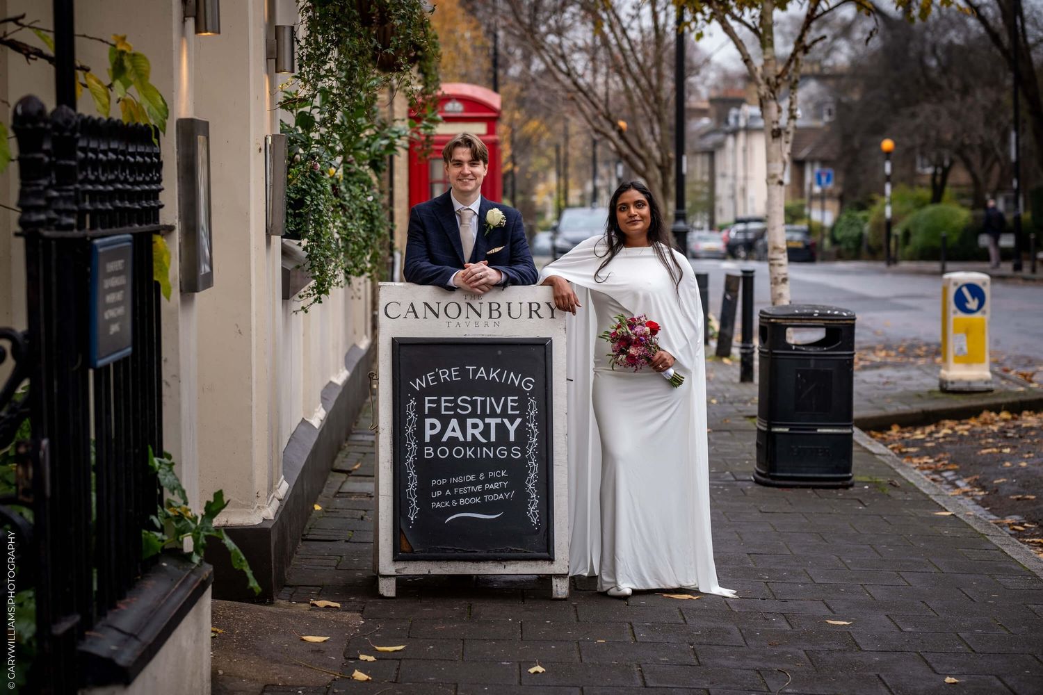 A couple in formal wedding attire stands next to a chalkboard sign outside Canonbury pub on a quiet street.