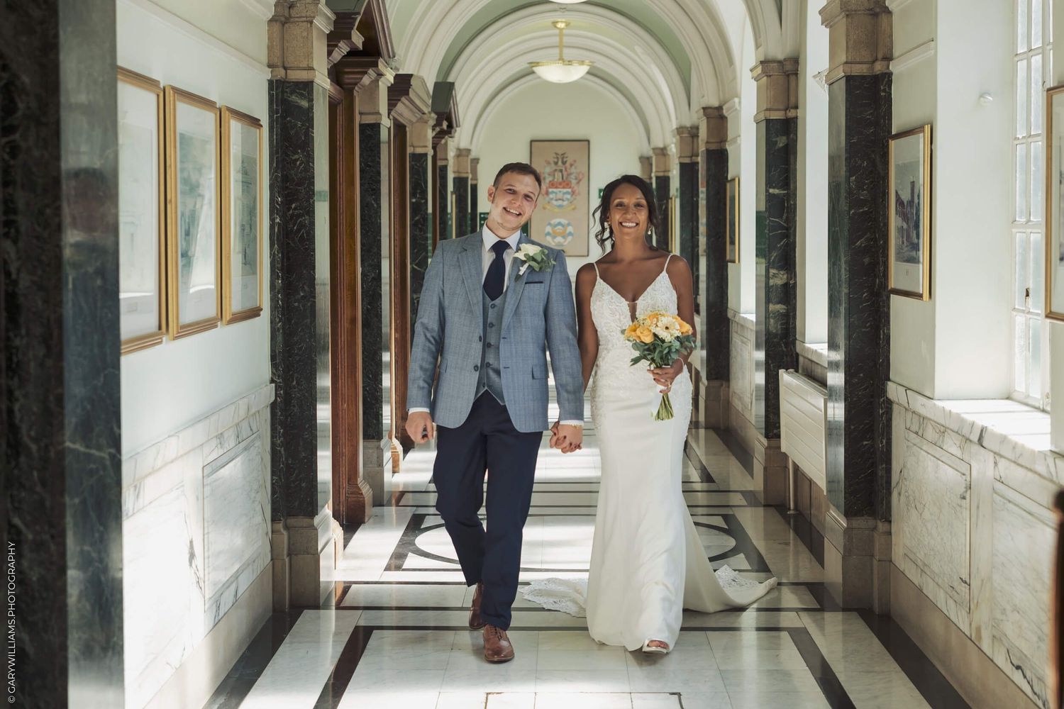 A bride and groom walk together down an elegant hallway with arched ceilings and framed artwork on the walls.